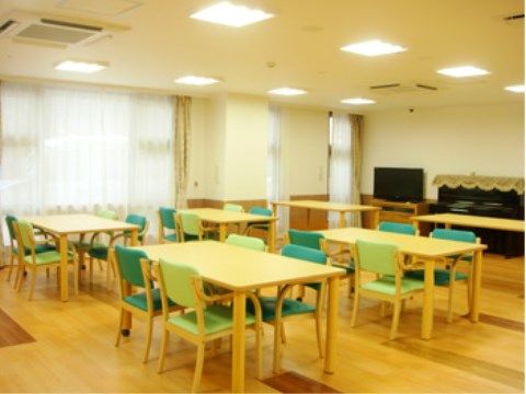 部屋の奥にテレビと、上部にカバーのかかったピアノが置かれている。左側には大きな窓が複数あり、レースのカーテンが引かれている。