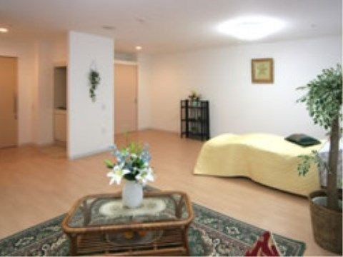 白い壁にフローリングの床の部屋である。広い室内に、ベッド、テーブル、観葉植物が配置されている。入り口近くには収納スペースが備わっている。