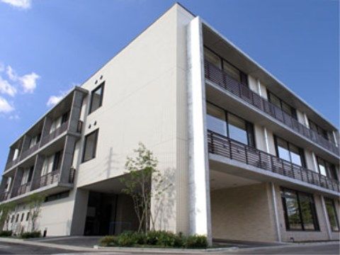 玄関前にある植え込みに背の高い樹木が生えたベージュの建物