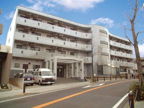 白い４階建ての建物で、玄関前には階段があり、柱が立っている。階段の左側に二台分の駐車スペースがある。
