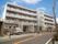 白い４階建ての建物で、玄関前には階段があり、柱が立っている。階段の左側に二台分の駐車スペースがある。