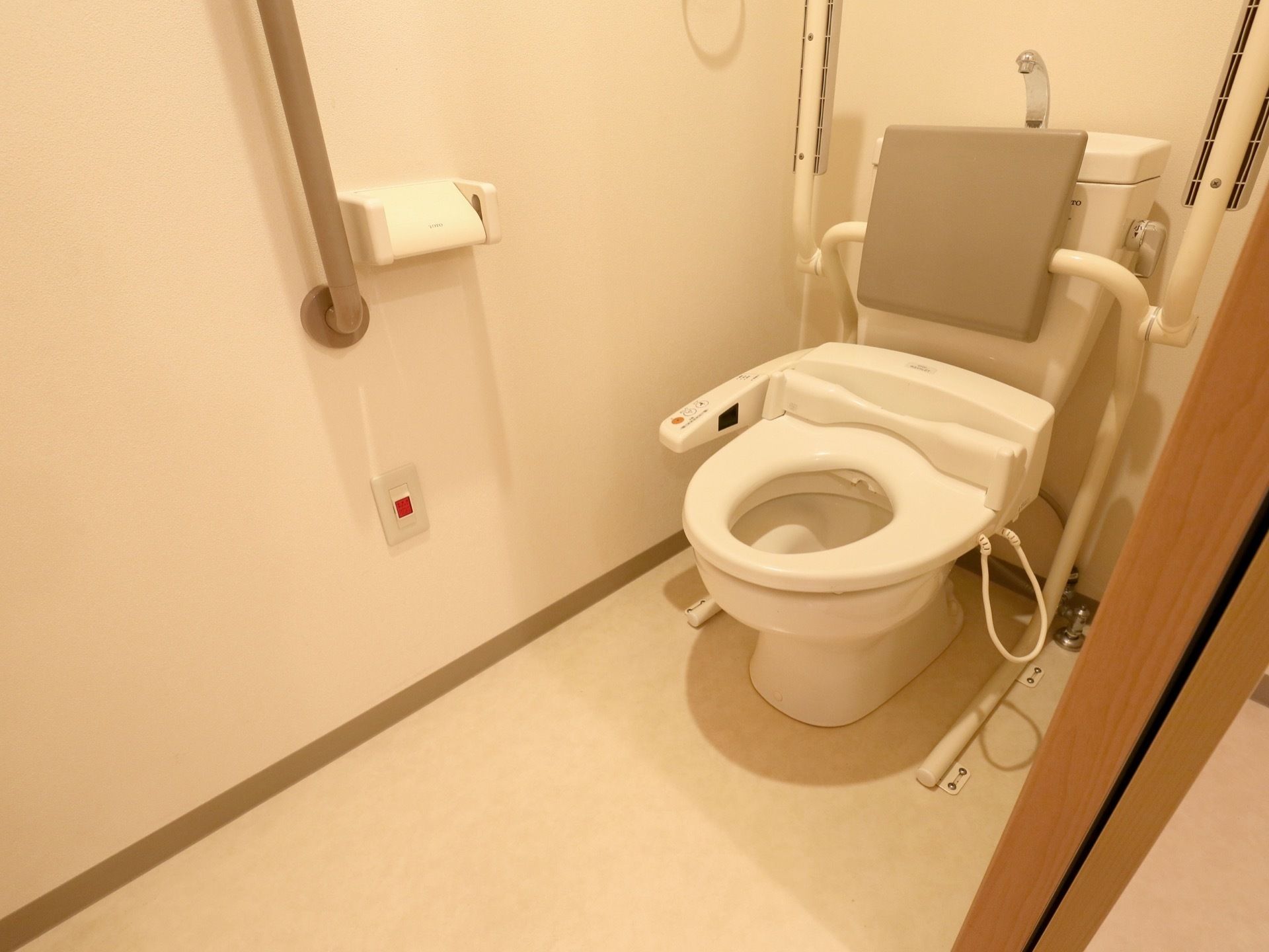 便座の両側に可動式の手すりがついた洋式トイレである。壁にもI字型の手すりがある。温水洗浄機能付きで、扉は引き戸になっている。