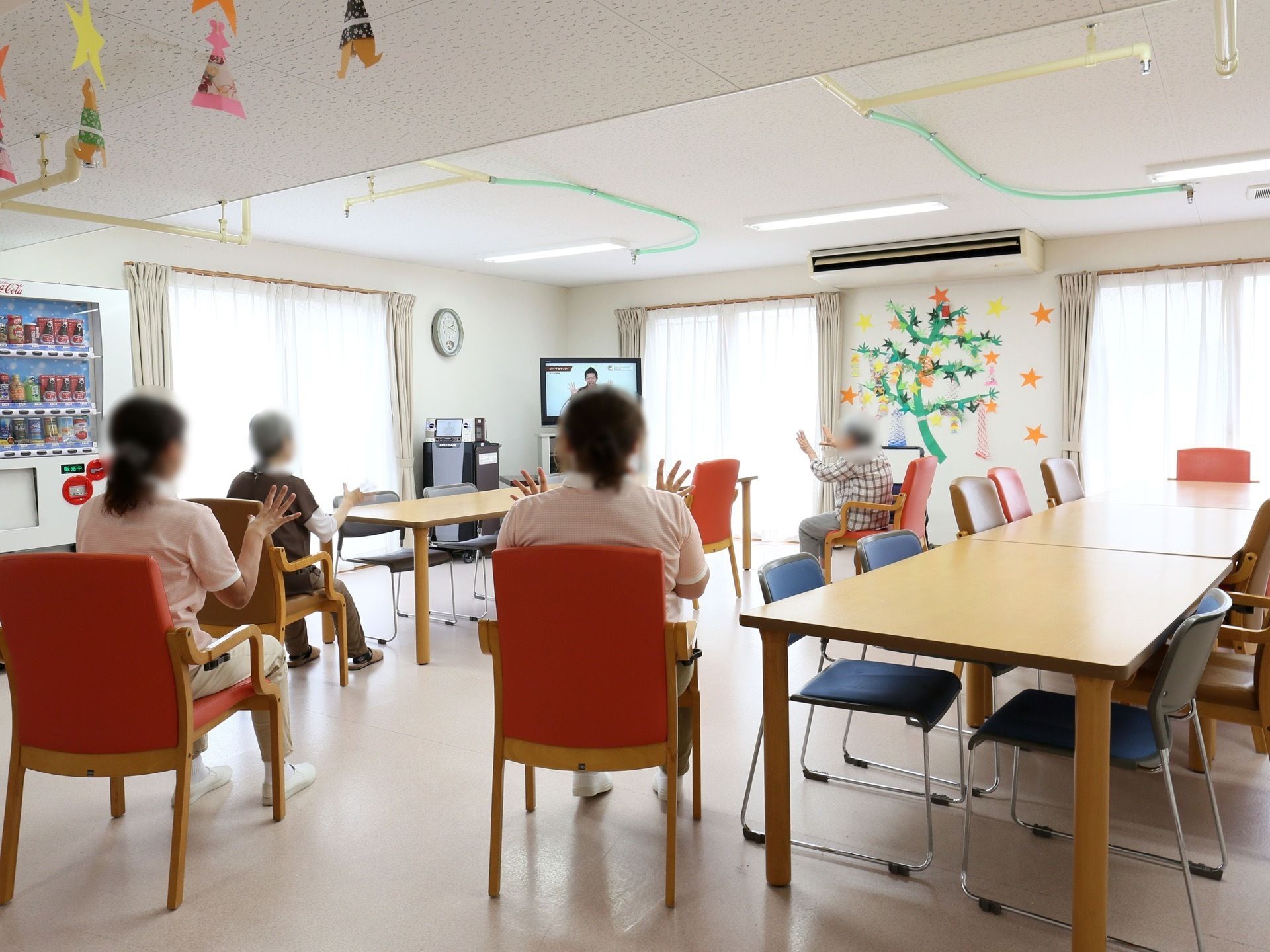 テーブルが並べられた共有スペースで、入居者様とスタッフが数名、テレビを見ながら体操を行っている。天井には空調が設置されている。