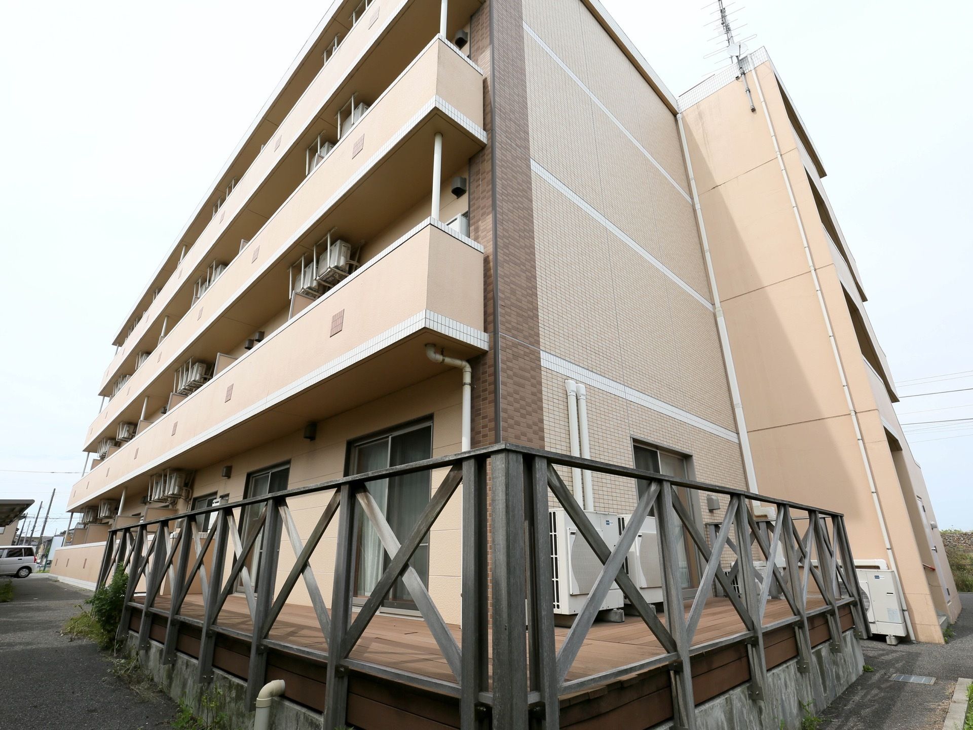 建物の敷地はアスファルトで舗装され、車が一台停まっている。1階の角にはL字型にウッドデッキが設置されている。