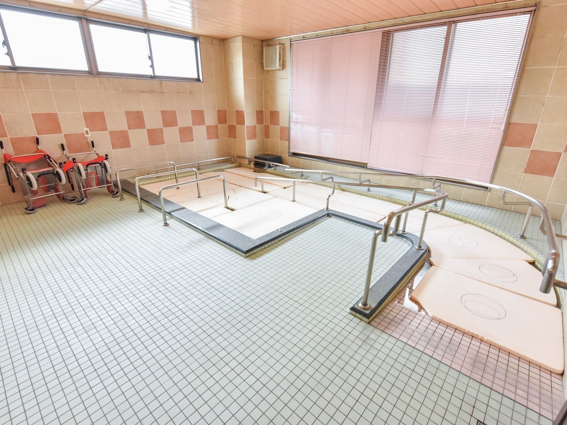 大浴場は大きな窓に面している。ブラインドが下げられている。入浴補助用のストレッチャーが２台置かれている。