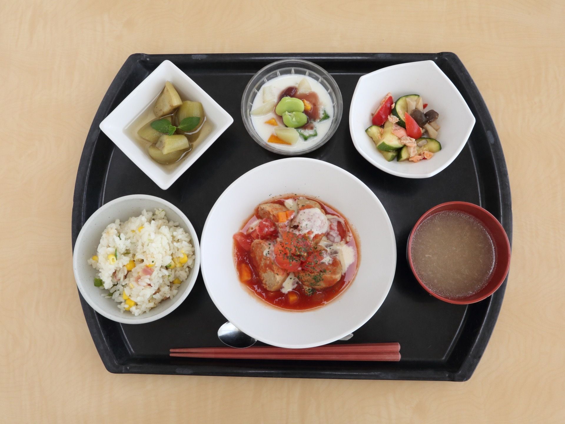黒いお盆の上に小さなお皿に盛りつけられたご飯やおかずが数多く並んでいる。手前には赤いお箸と小さなスプーンがある。