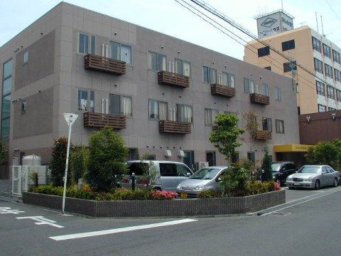 交差点の角にある、植込みに囲まれた駐車場とその奥に建つライトブラウンの建物