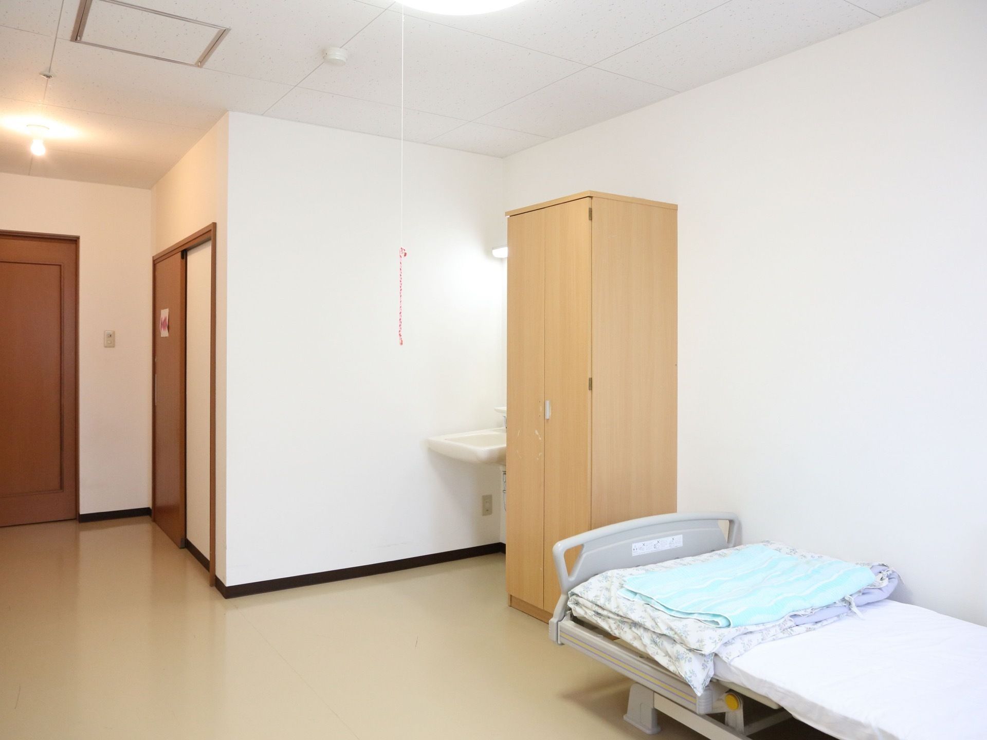 電動ベッド、洗面台完備である。収納スペースとしてタンスが設置されている。白を基調とした、あたたかみのある部屋である。