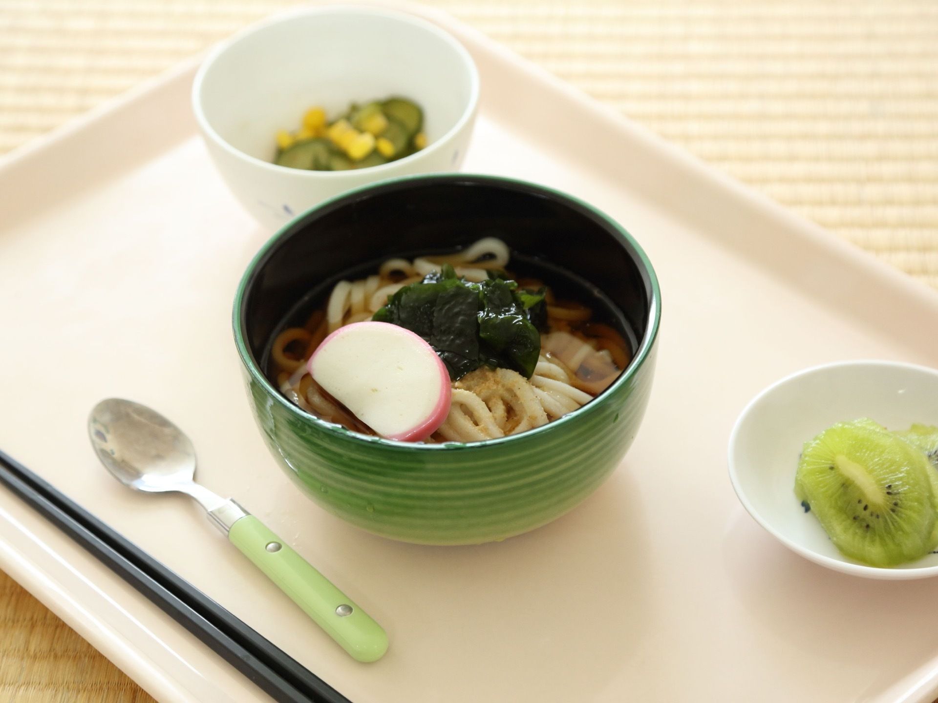 トレーに、お箸、スプーン、かまぼことわかめが入ったうどん、きゅうりとコーンのお浸し、キウイフルーツがある。