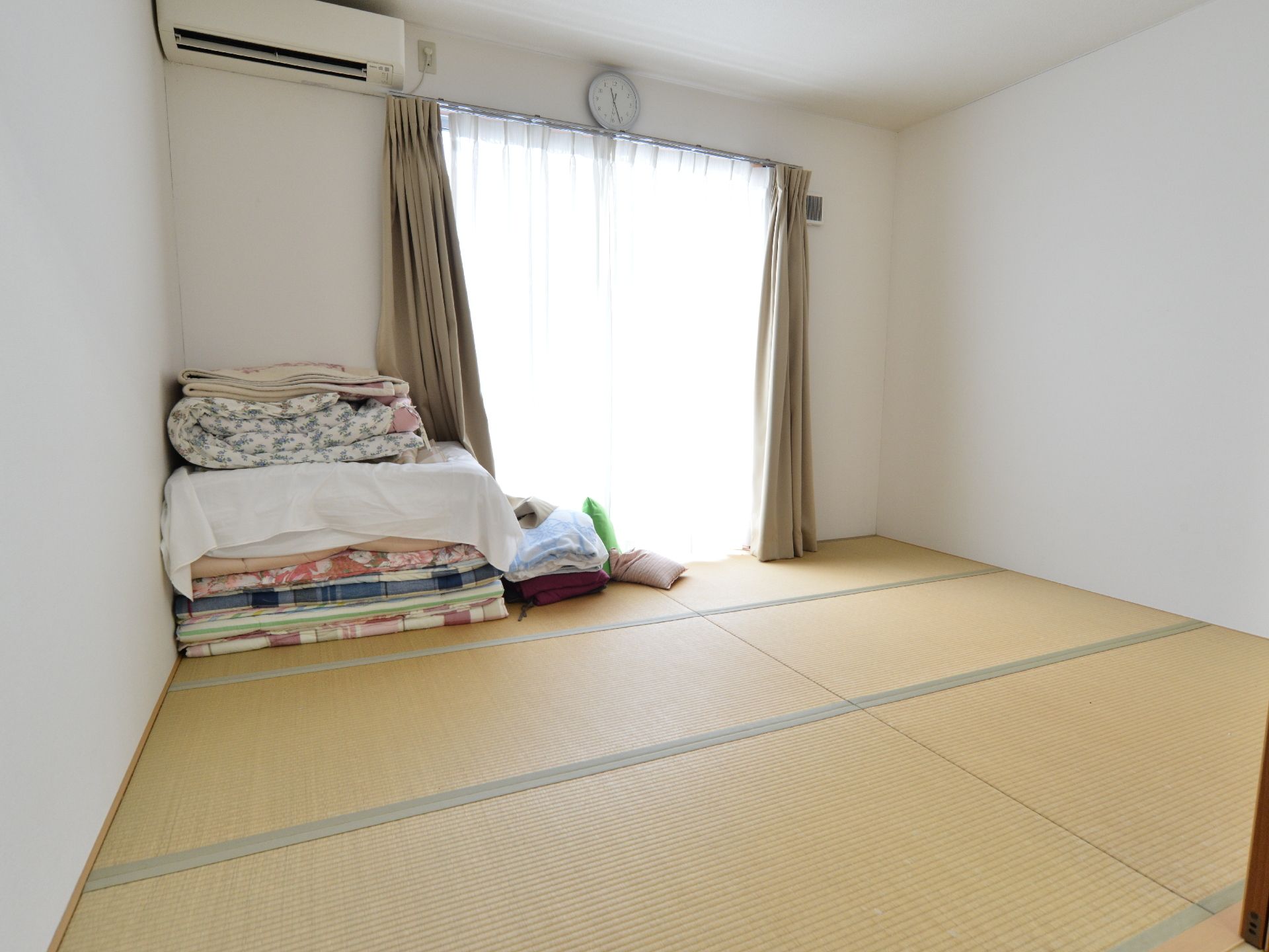 和室の部屋。布団が畳まれて部屋の隅っこに置かれている。壁にエアコンとアナログの時計が設置されている。
