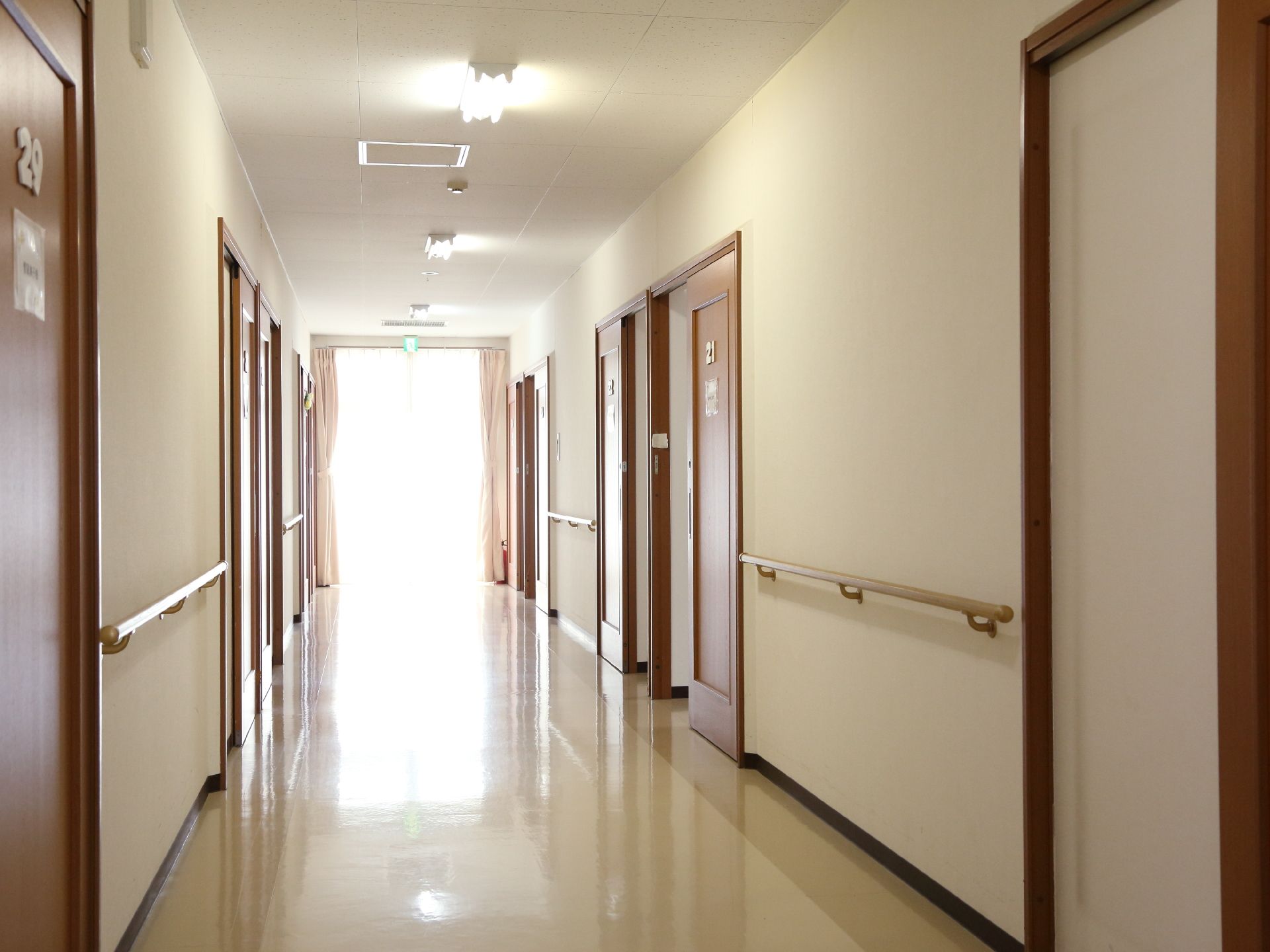 突き当りに掃き出し窓がある通路で左右の壁に手すりが設けてある。誘導灯が奥に見える。通路の両サイドに部屋がある。