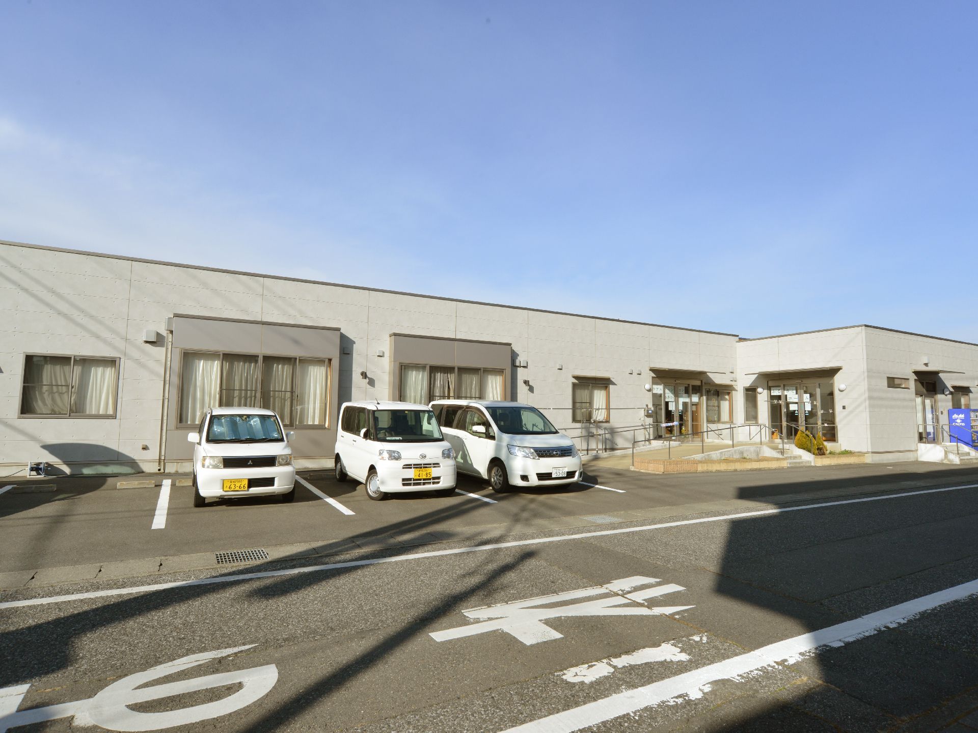 グレーの平らな建物になっており、右側に入り口がある。目の前は駐車場になっており車が何台か停まっている。