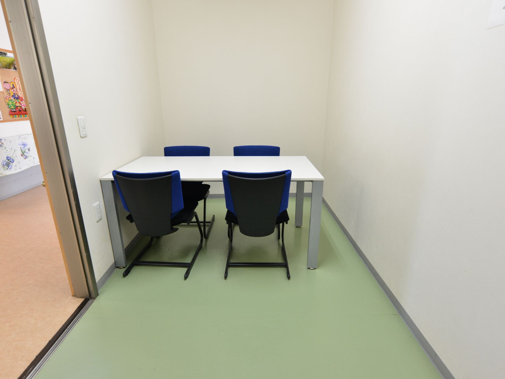 白壁と緑色のフローリングの床の部屋に白いテーブルと黒い椅子が4脚用意されている。左手は廊下になっている。