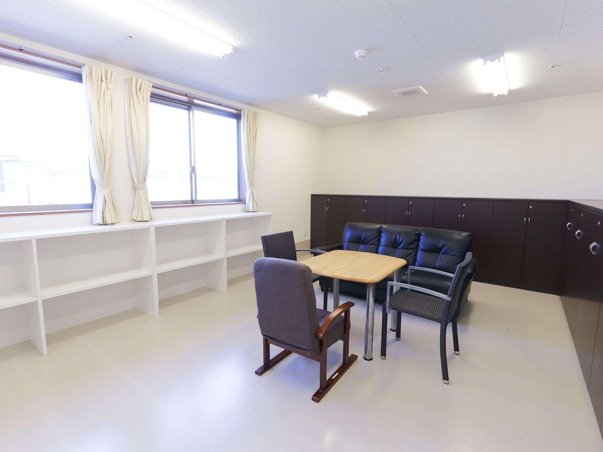 窓が2つあり、明るい部屋になっている。テーブルや椅子、ソファを設置している。壁には棚や収納などを設置している。