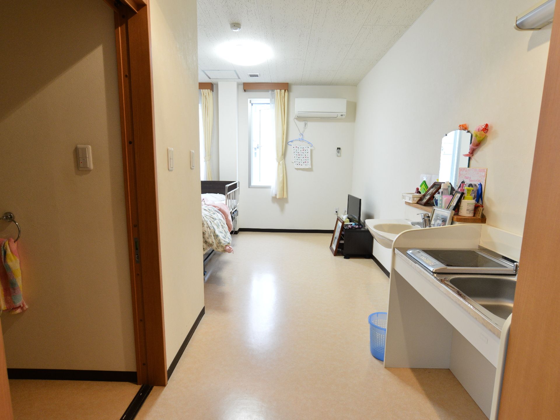 白色の天井と壁にクリーム色の床の内装。右手にミニキッチンが設置され、隣には洗面台がある。奥に進めば介護用ベッドがあり、正面の壁にはエアコン。