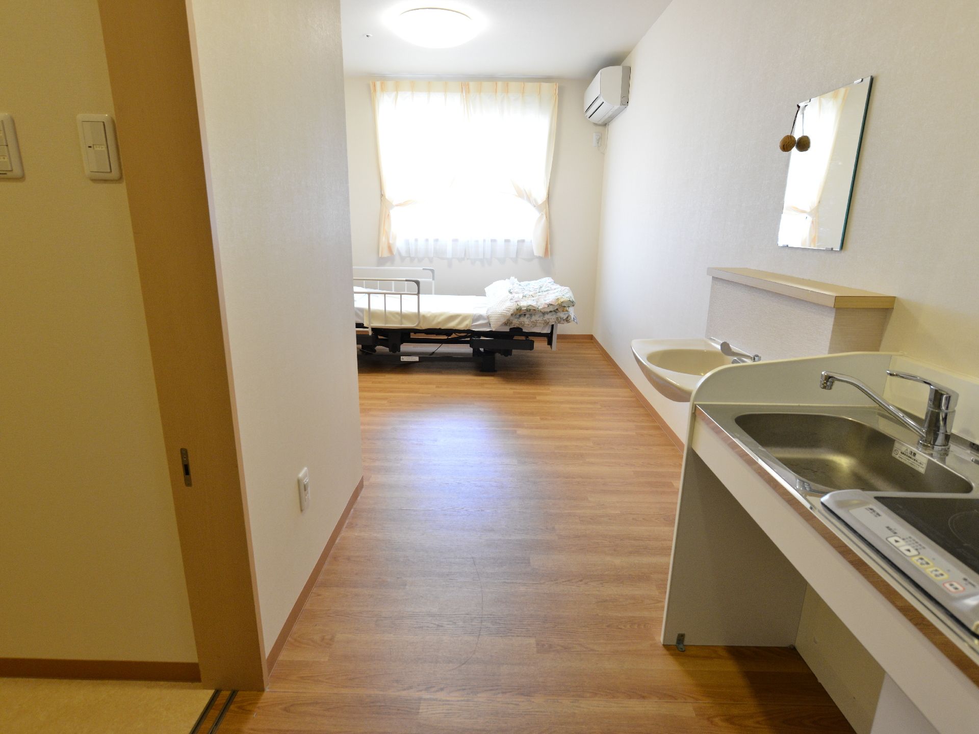 居室には、1口のIHコンロと流し台、洗面所がある。窓際にはベッドが設置されている。またエアコンも取り付けてある。