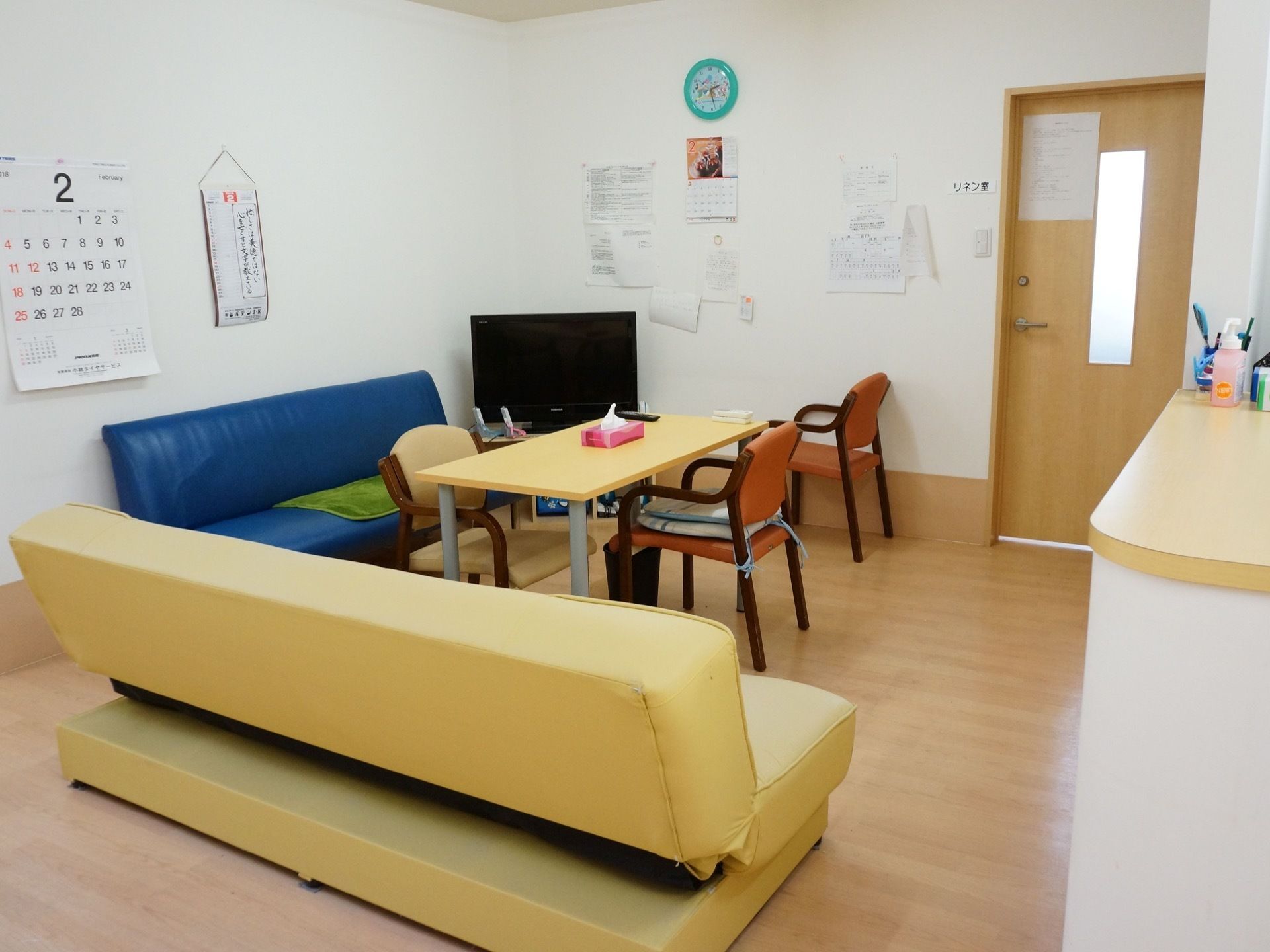 部屋の隅にテレビが置かれ、その前に机と椅子、ソファが並べられている。椅子の上には座布団が置かれている。