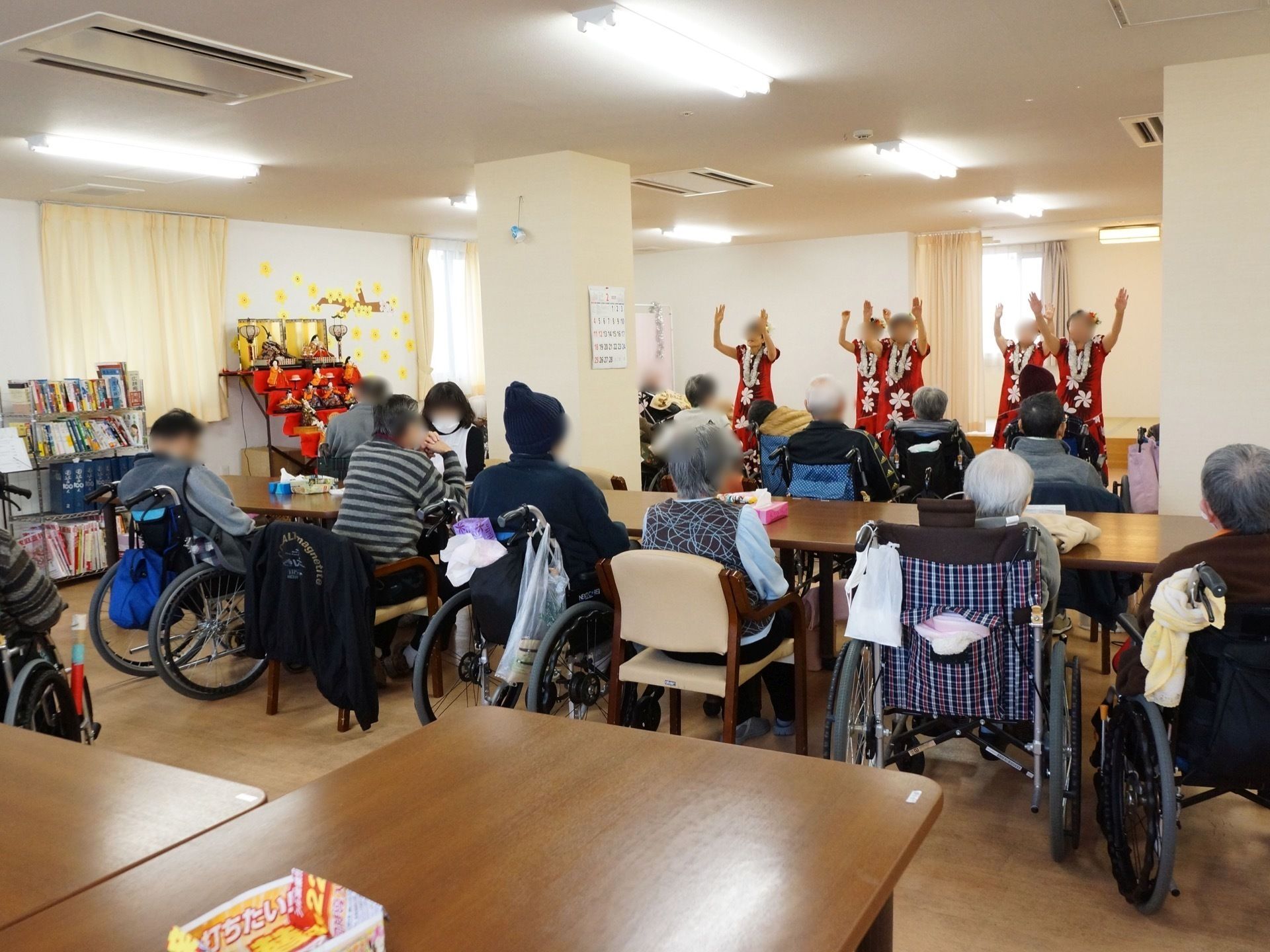 椅子や車椅子に座ったり入居者様方が、踊りの催し物を楽しんでいる。壁際にはひな人形が飾られ、季節を感じることができる。