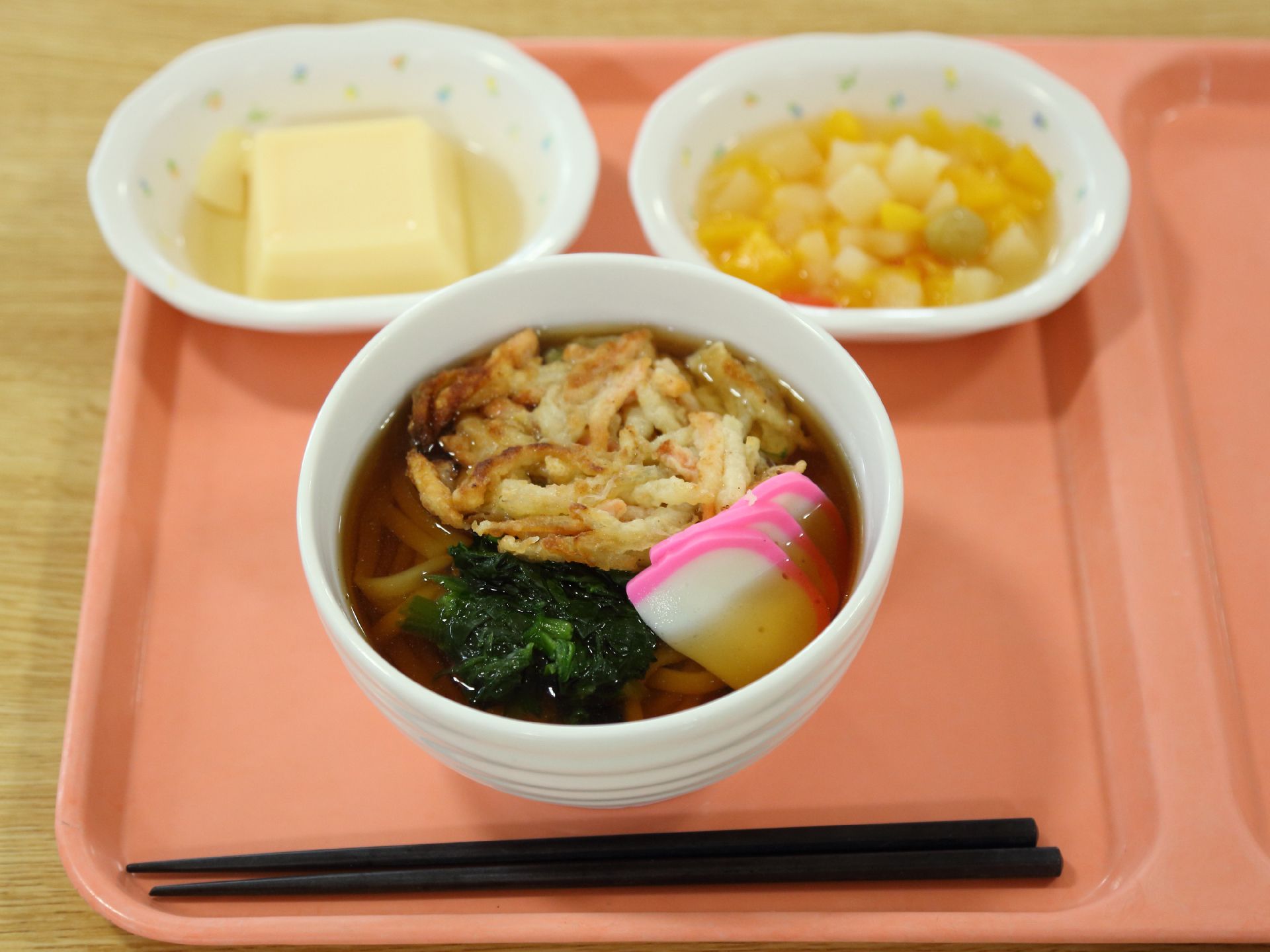 施設内で提供される、食事の一例。ピンクのトレーに箸が置かれ、食事が並んでいる。うどんとおかず、デザートがある。