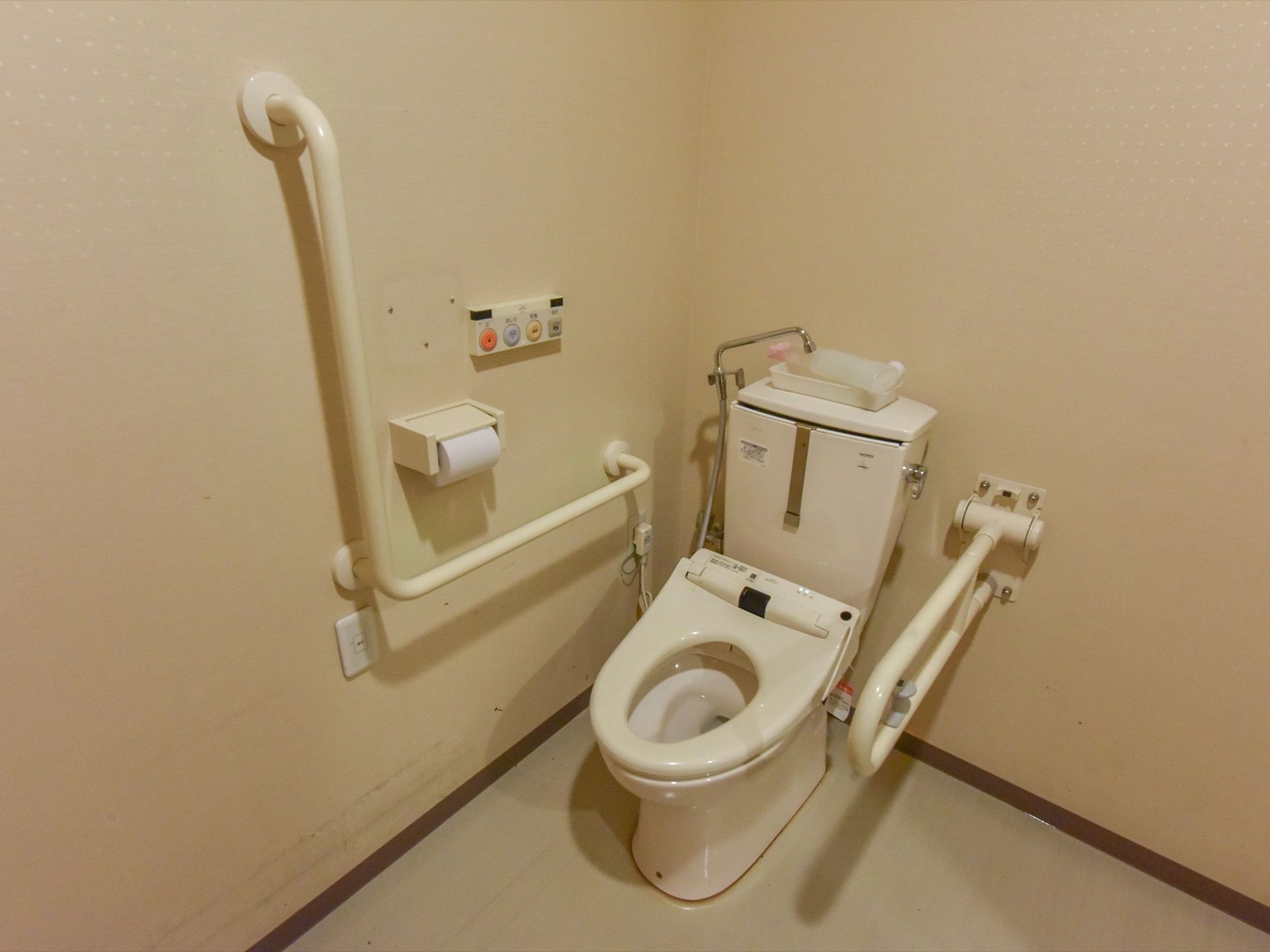 トイレは洋式で、洗浄機能がついている。白くしっかりとした造りの手すりが取り付けられている。便器の周囲にはスペースが取られている。