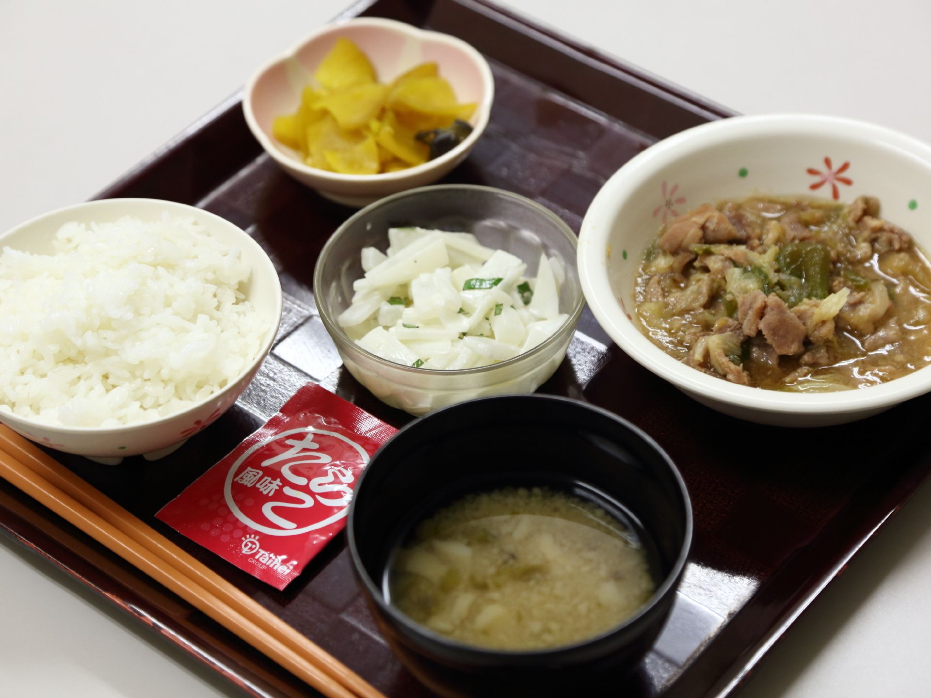 トレイに今日の食事が盛り付けてある。ごはん、味噌汁、ふりかけがある。細かく切った野菜と肉の煮物。その他小鉢が二つついている。