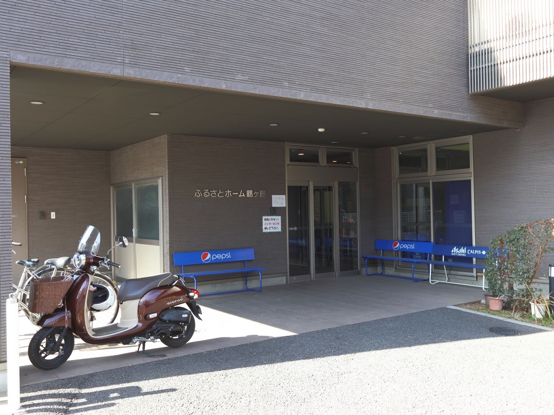 玄関の外観である。１階が削られた形になった部分にある出入口があり、車をすぐ前に停められる。玄関口には青いベンチが３脚置かれている。