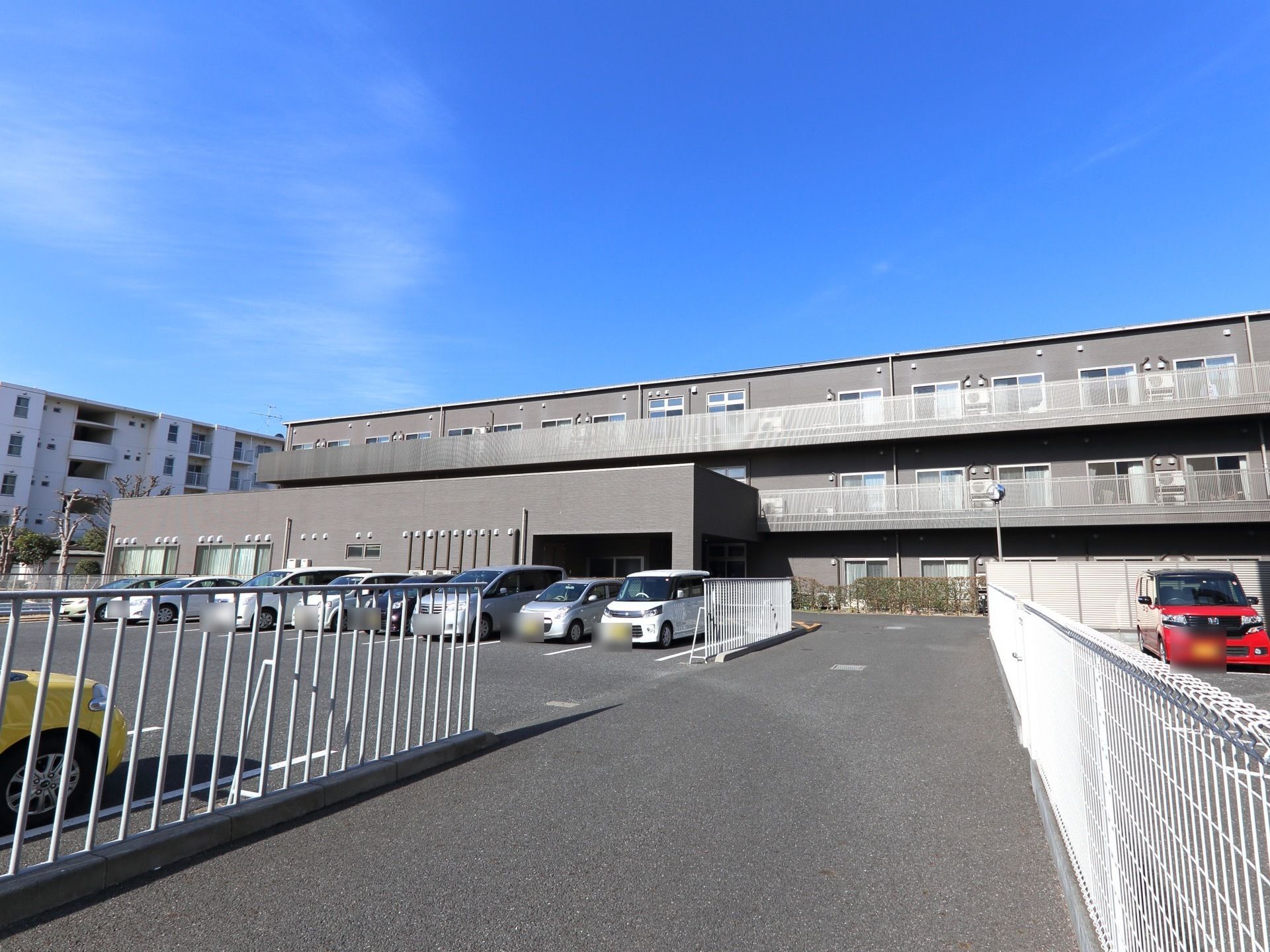 施設はグレーの外壁の３階建てで、横に細長い建物である。敷地が非常に広く大きな駐車場があり、たくさんの車が停められている。