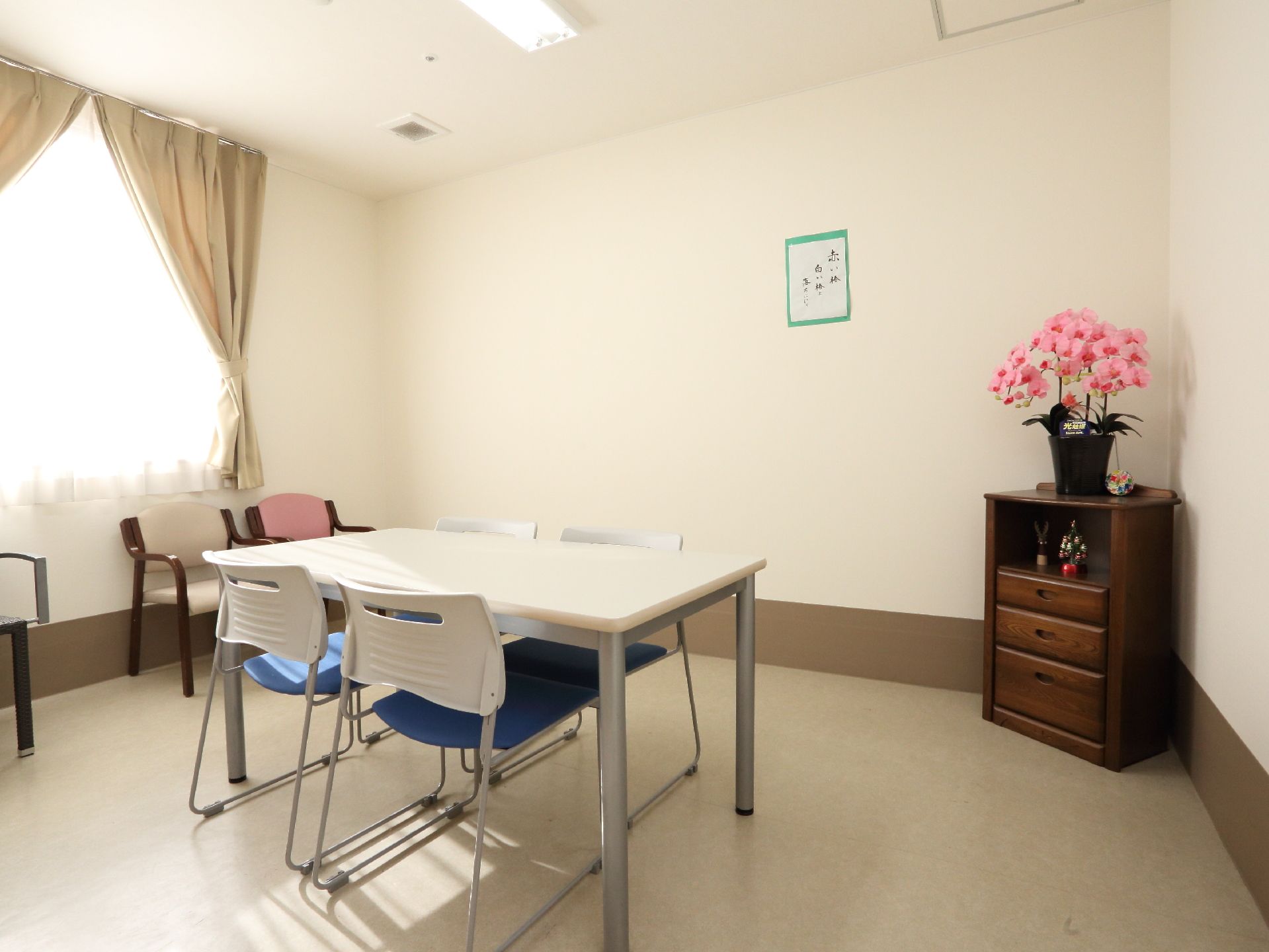 部屋の中央に机と椅子があり、窓側には椅子のみ置かれている。壁には書道が飾られている。木製のタンスの上には美しい花がある。