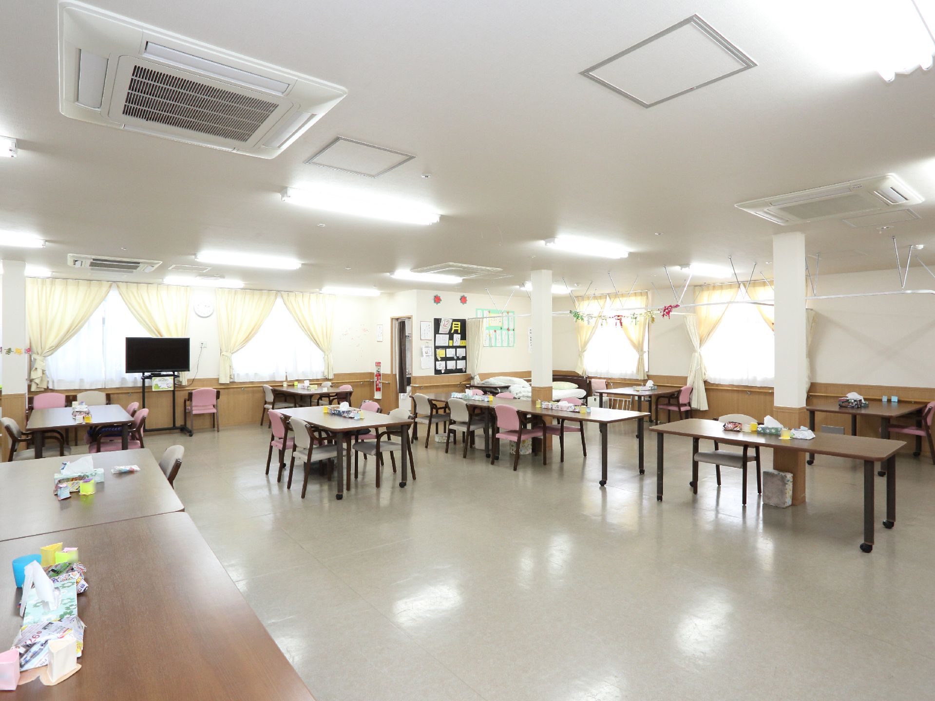 広々とした食堂に複数の椅子とテーブルが置かれている。窓際にはテレビがある。天井にはエアコンが設置されている。