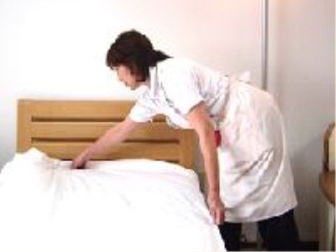 木調デザインのベッドのシーツを整えるエプロン姿の女性