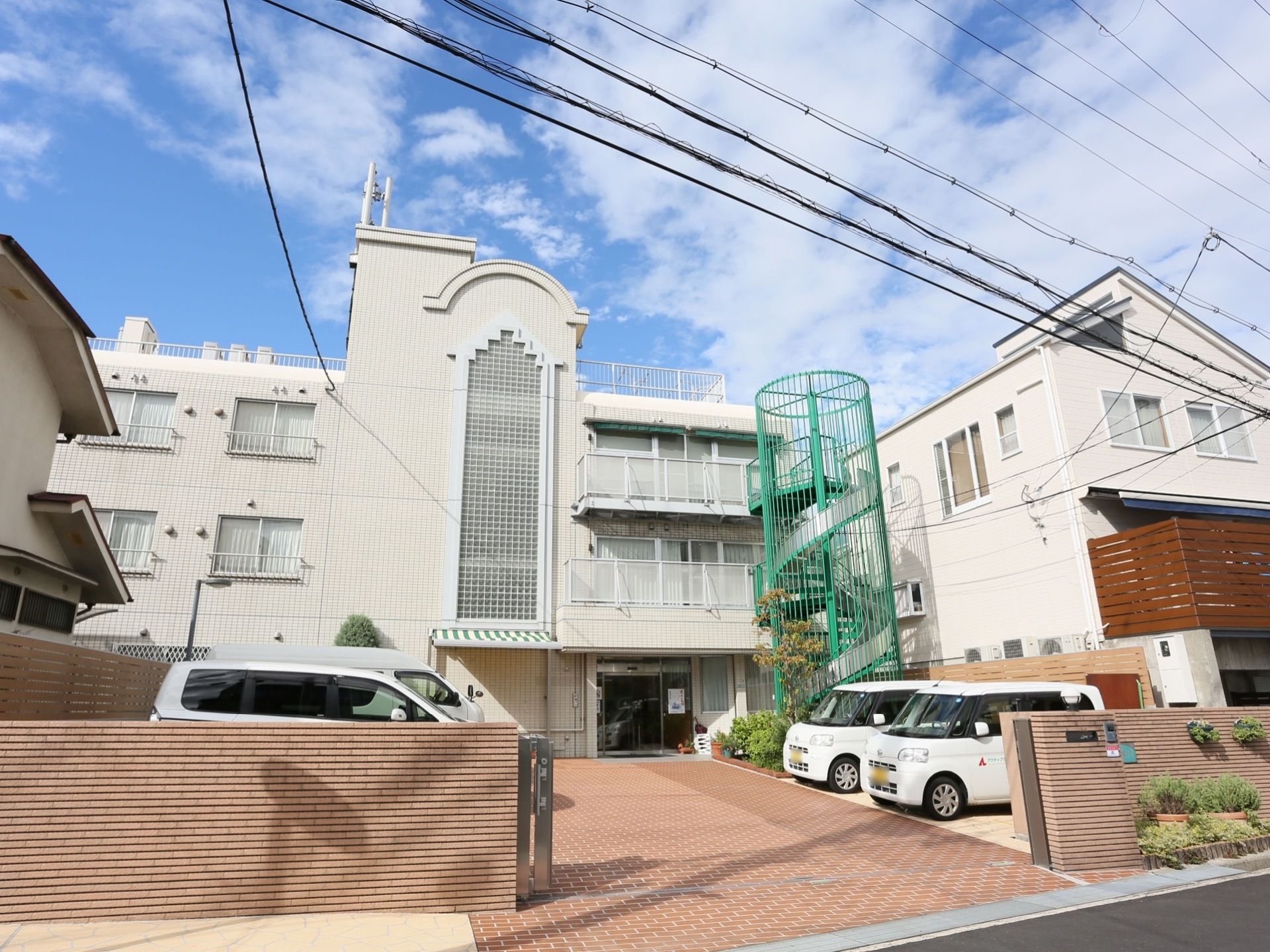 白色を基調とした3階建ての施設の建物に、緑色のらせん階段が備わった外観である。敷地内には駐車スペースが設けられている。