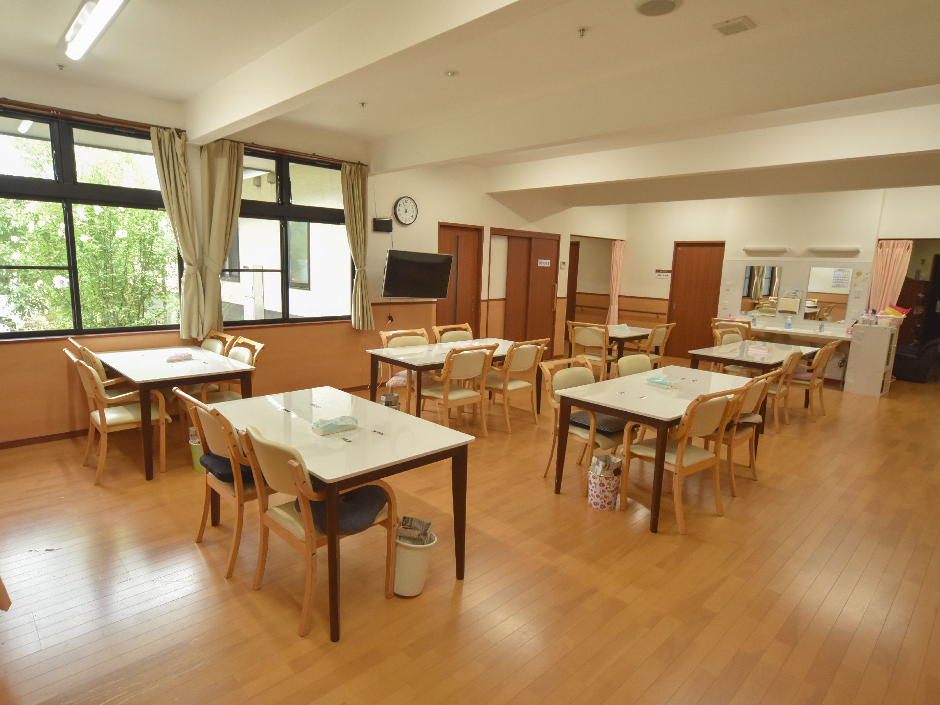 天井が高く、広々とした食堂である。右手には居室のドアがある。壁に時計とテレビがかけられている。各テーブルにティッシュとゴミ箱が置いてある。