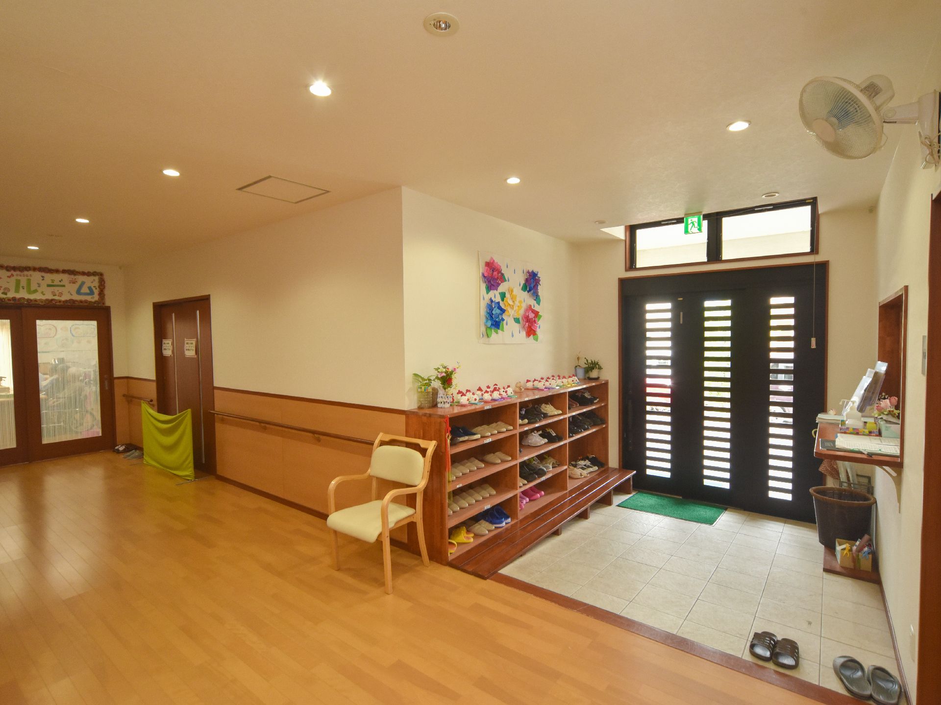 玄関の靴用の棚にスリッパや靴が並んでいる。ドアからスロープで館内に入ることができる。左奥にデイルームがある。