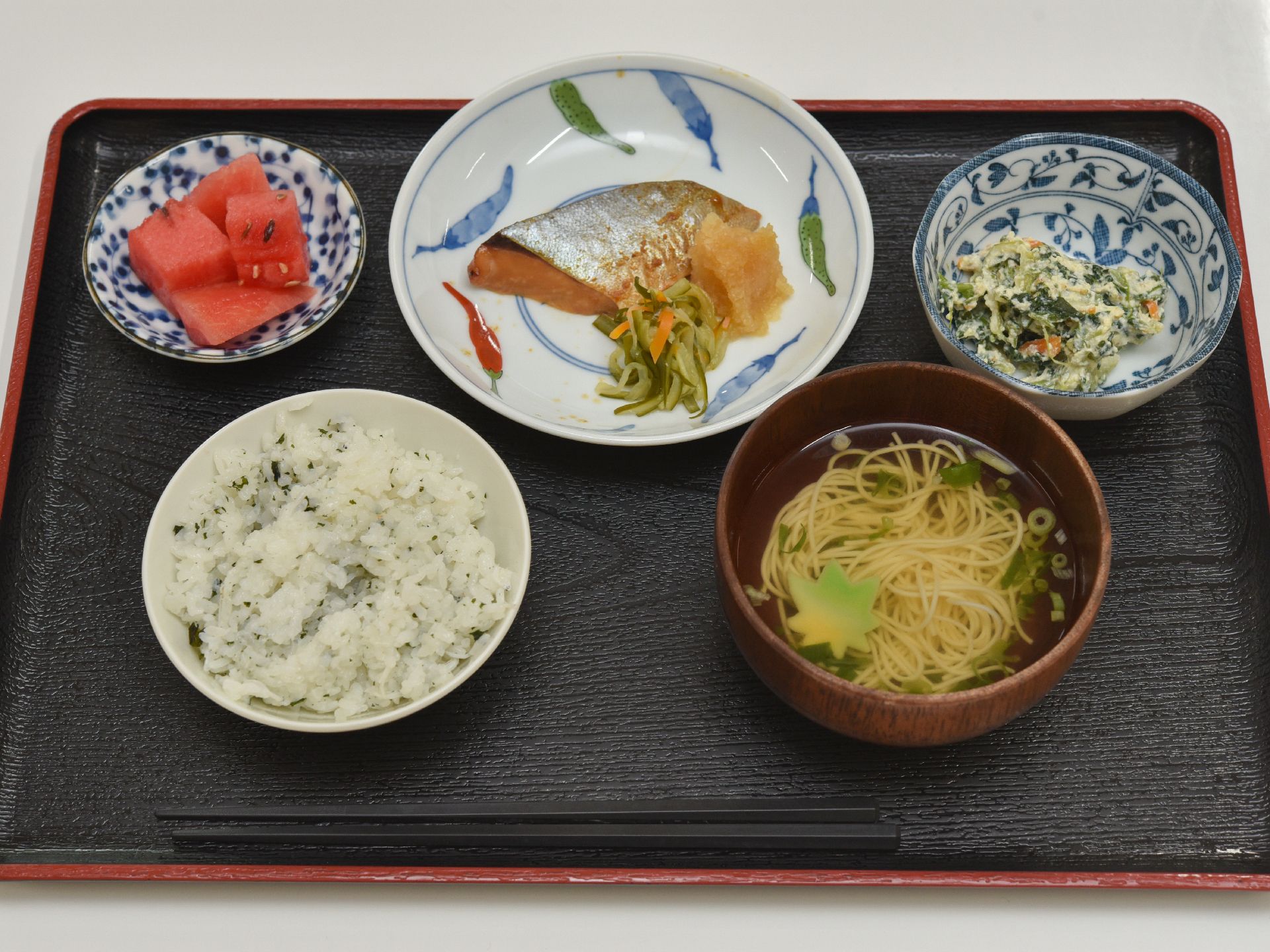 わかめご飯、そうめんの入ったおつゆ、主菜の魚に付け合わせ、白和え、デザートのスイカがトレーに載っている。