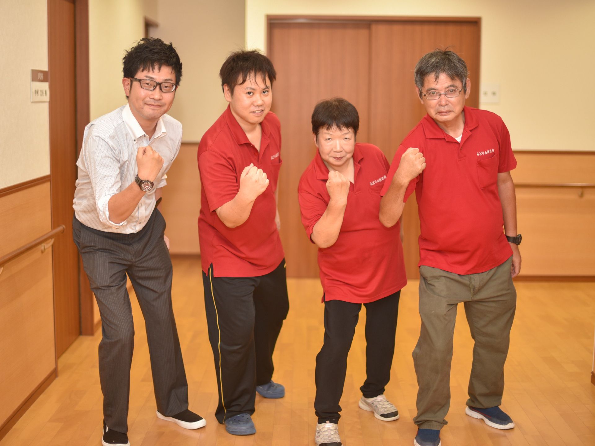 施設の廊下で、そろって右手の拳を出したポーズをとる４人のスタッフ