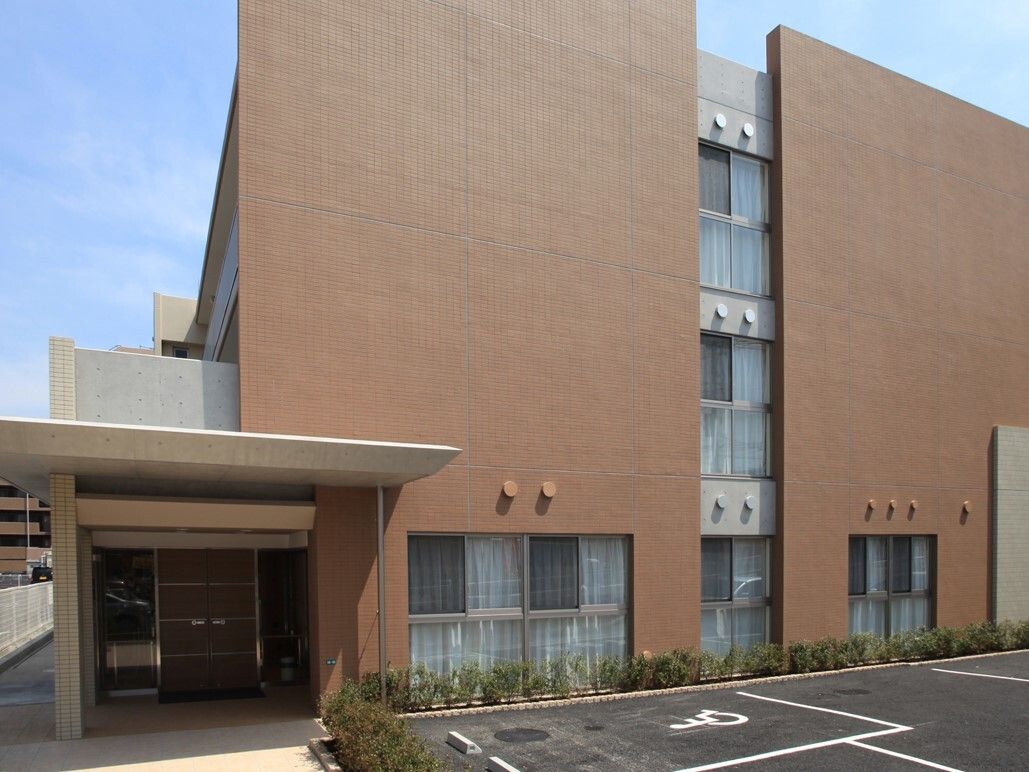 ３階建ての建物を覆うように茶色い壁が２枚立っている。敷地内には駐車場があり、歩行者用通路は区別して確保されている。