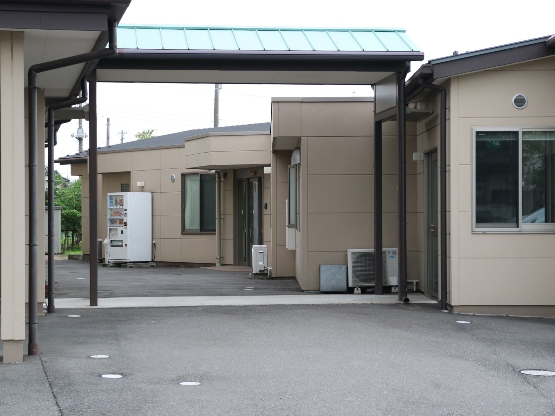 施設と施設をつなぐ通路には緑色の屋根が付いている。周囲の地面はアスファルトで舗装されており、近くには飲み物の自動販売機がある。