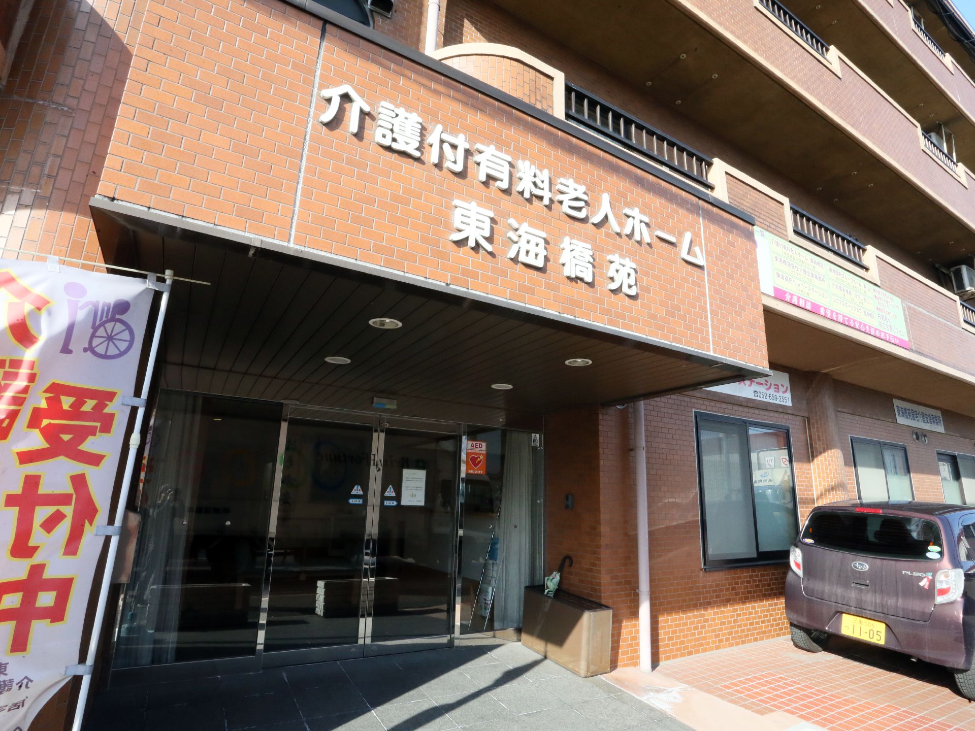 施設の外観は茶色いマンションタイプの造りになっている。入口には施設名が書いてあり介護受付中の旗が立っている。