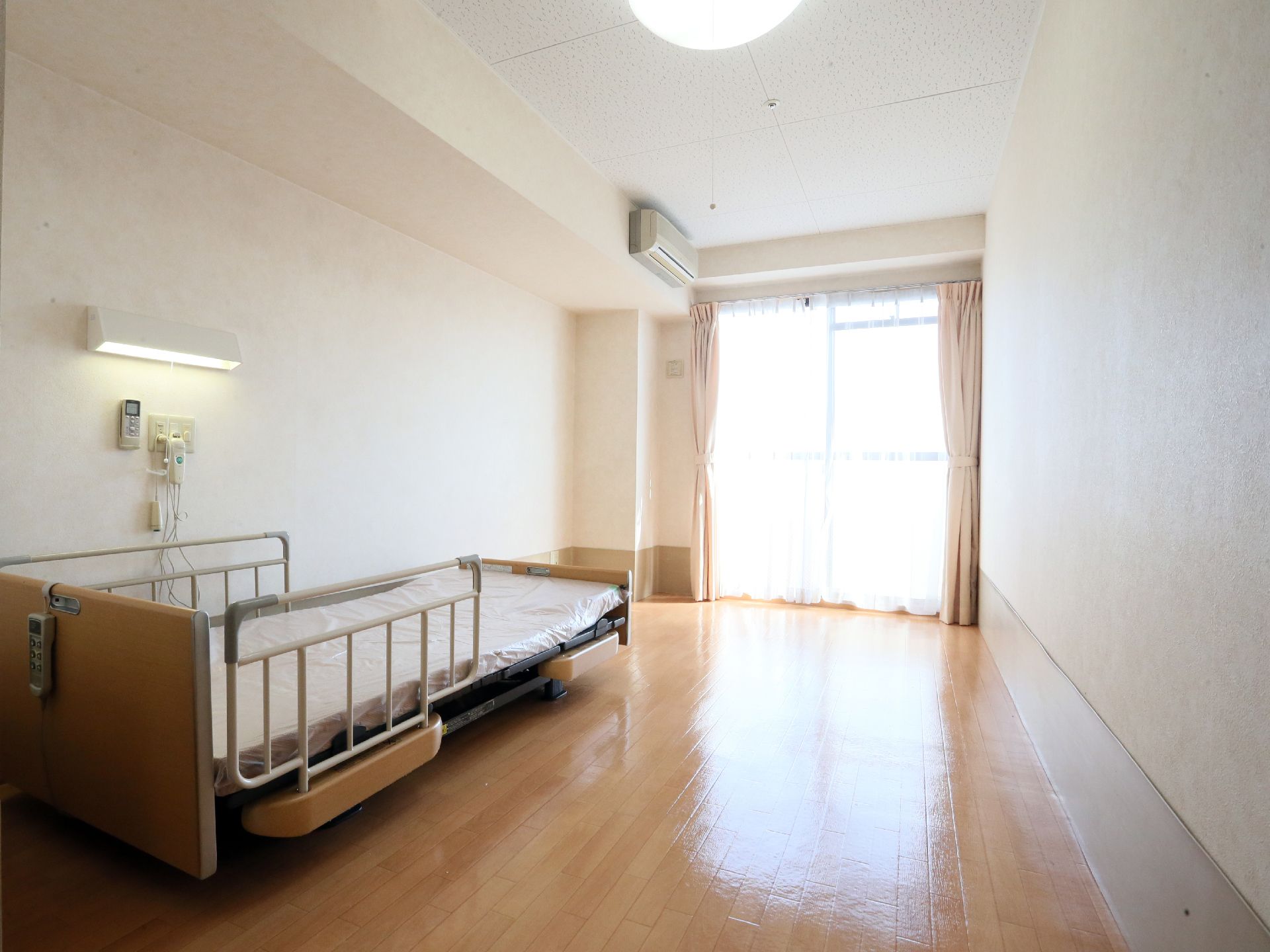 大きな掃き出し窓がある室内で、介護ベッドが置かれている。ベッド脇の壁には緊急呼び出しブザーや蛍光灯、エアコンのリモコンが設置されている。