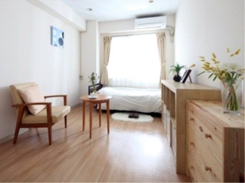 突き当りに腰高窓がある。上にエアコンがあり、窓下にベッドが置いてある。手前右側には整理ダンスと、間を空けて一人掛けの椅子がある。