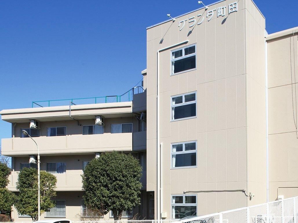 外壁が白い４階建ての建物で、居室窓の前にはバルコニーが付いている。２棟がＬ字型に連結しており、一方は屋上に柵がある。