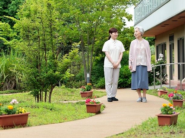 戸外で日に当たる白髪の車椅子利用者と近くのベンチに座る女性