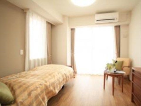 エアコンが完備された日当たりの良い個室タイプの居室である。ベットや丸テーブル、椅子などの家具が置かれている。