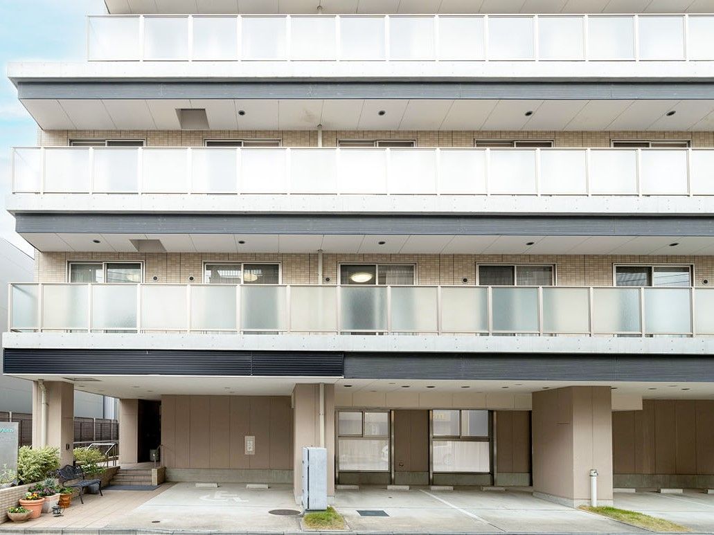 団地のような見た目の４階建ての建物。１階部分は駐車スペースになっている。周辺には民家やマンションなどがある。