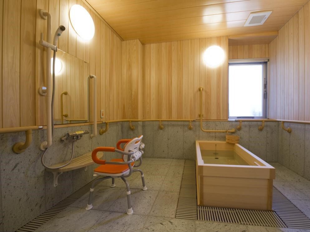 木製の浴槽を備えた、広々とした浴室である。壁にも木が使われており、手すりが備わっている。洗い場に椅子が置かれている。