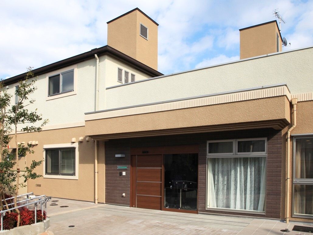 白とアイボリーのツートンカラーの２階建ての建物である。建物前には木々が多く植えられている。玄関は茶色のドアである。