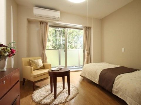 居室の窓からは日が差し込み、室内が明るくなっている。ベッド、テーブル、椅子、タンスが置かれている。タンスの上には花が飾られている。
