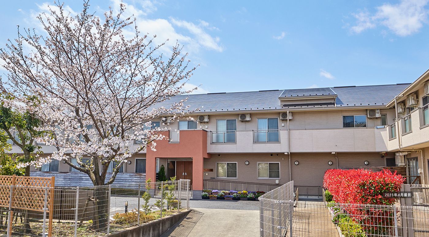 ベージュ色の外観で二階建ての建物は、正面扉が木製の自動ドアになっている。玄関までは手すり付きの階段か、スロープを利用する。