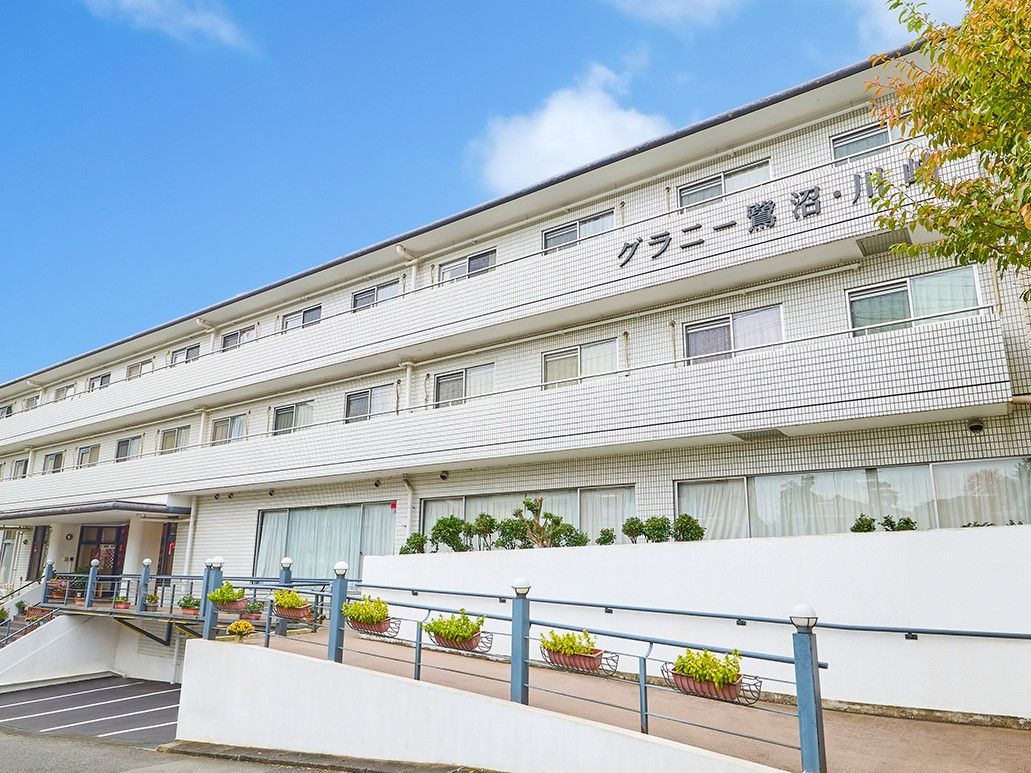 ３階建ての、明るいグレーの建物である。建物の近くには背の高い木があり、フェンスにはたくさんの花が飾られている。