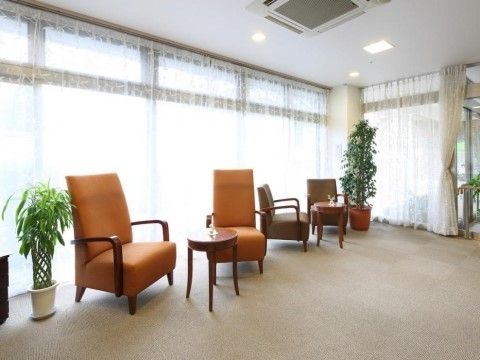 茶色とオレンジ色の一人用ソファーが４脚、小さな丸テーブルを囲んでいる。大きな窓には目隠しになるレースカーテンがかかっている。